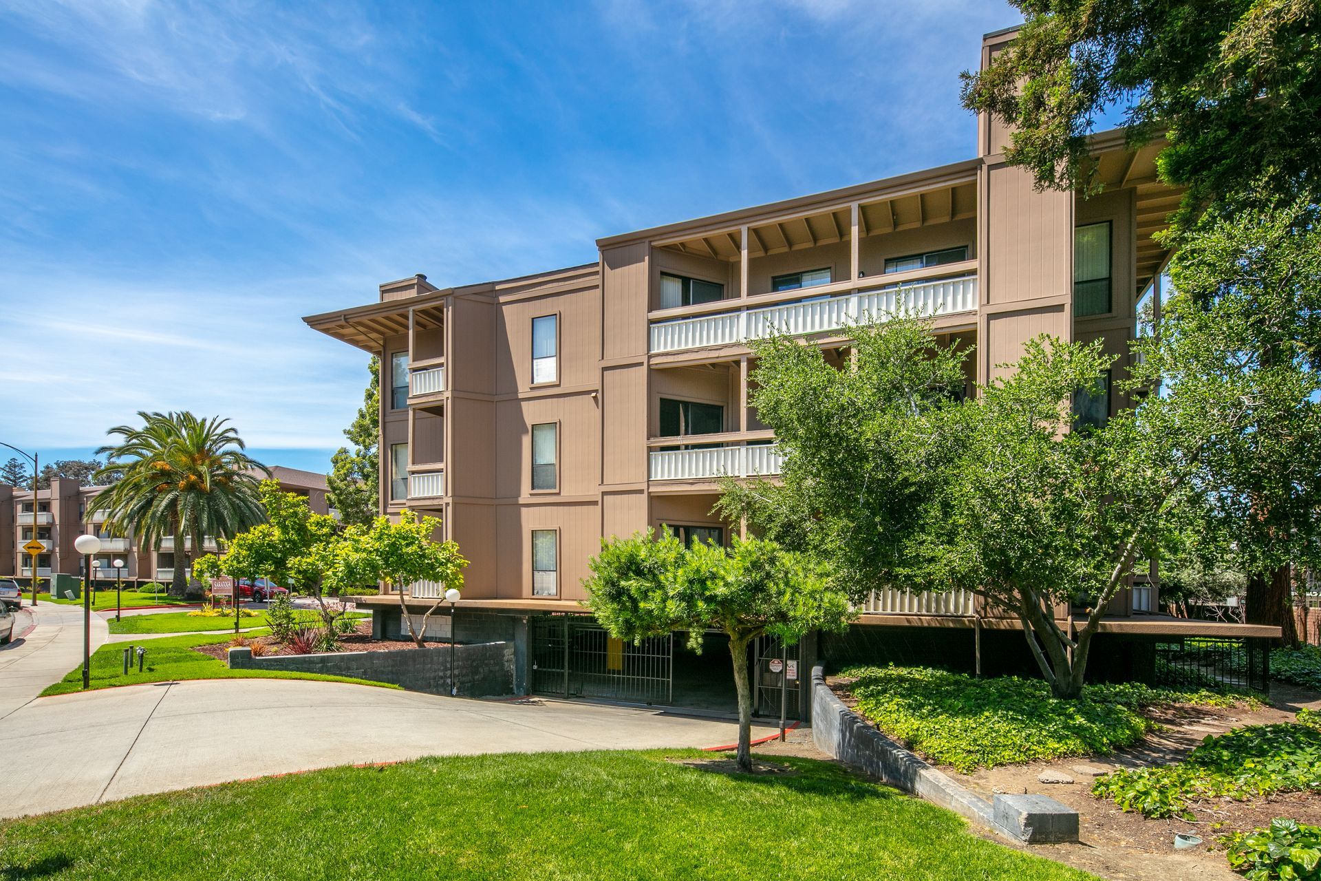 Modern apartment building with balconies, trees, and a green courtyard under a blue sky.