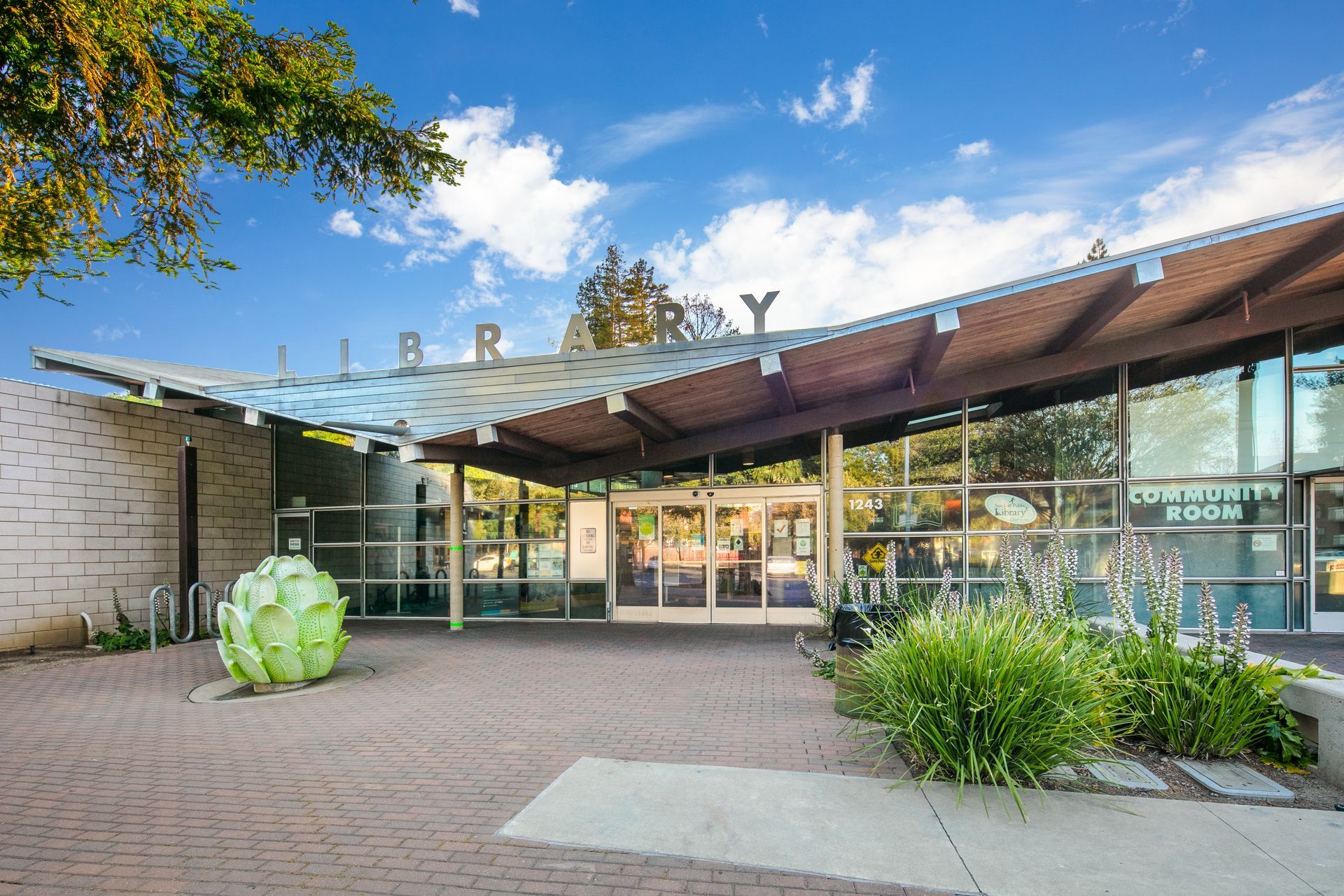 Modern building entrance with glass facade, curved roof, and landscaped courtyard under blue sky.