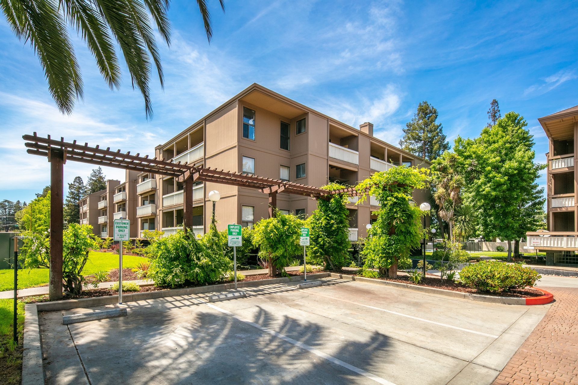 Apartment complex entrance with lush greenery, pergola walkway, and sunny blue sky.
