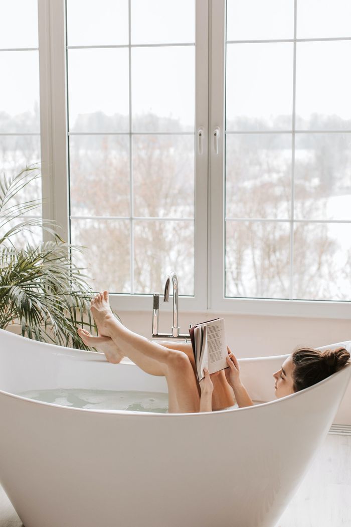 Woman  soaking an reading in bath tub next to large white aluminum framed casement window