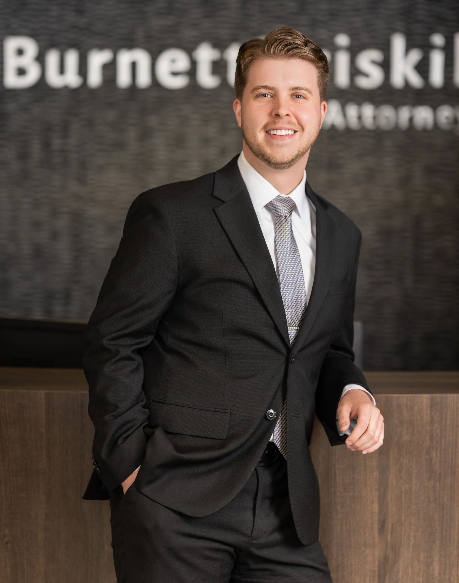 Man in suit smiling, leaning against a desk. Behind him is a law firm's name.