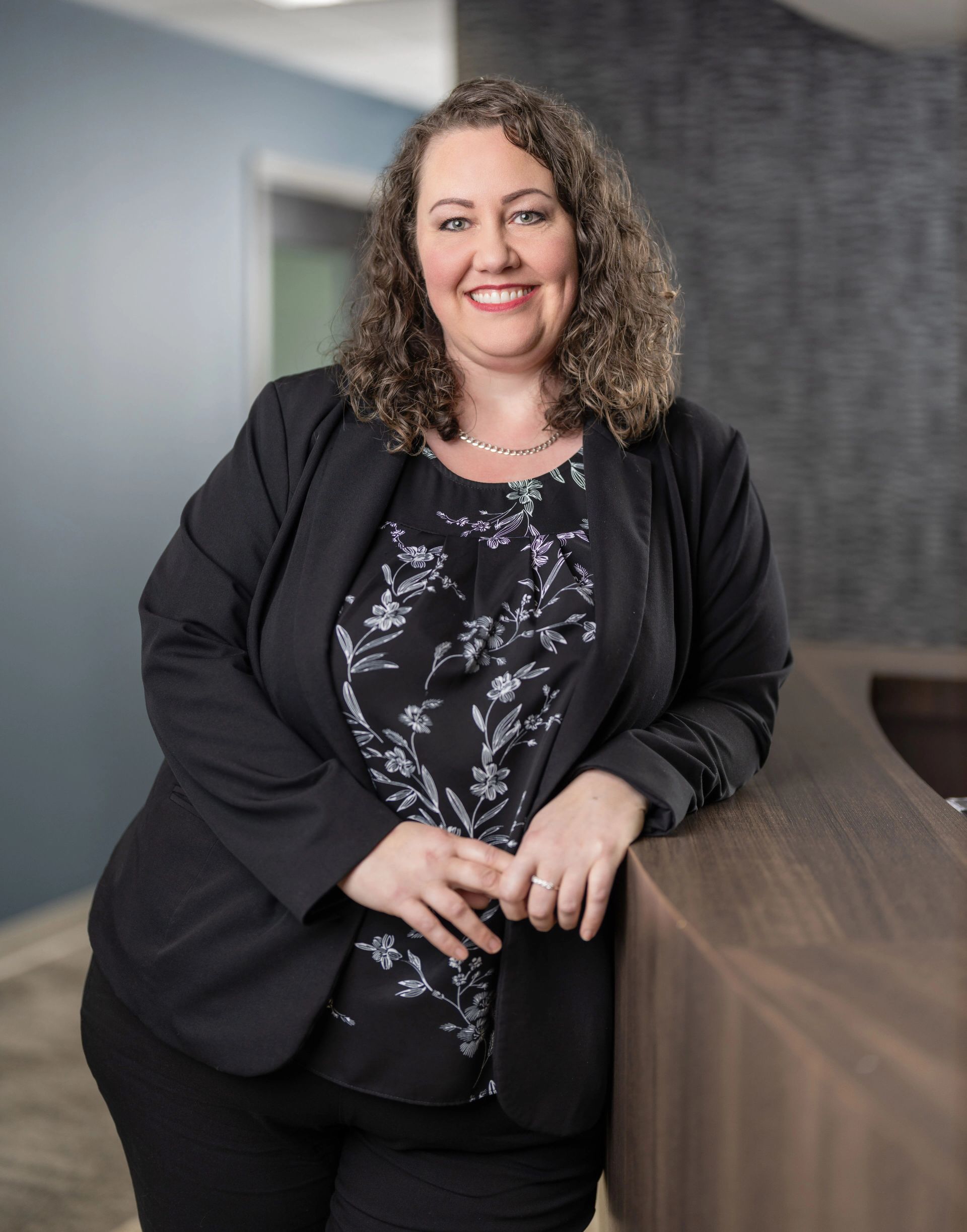 Woman in blazer smiles, leaning on a counter, in an office setting.