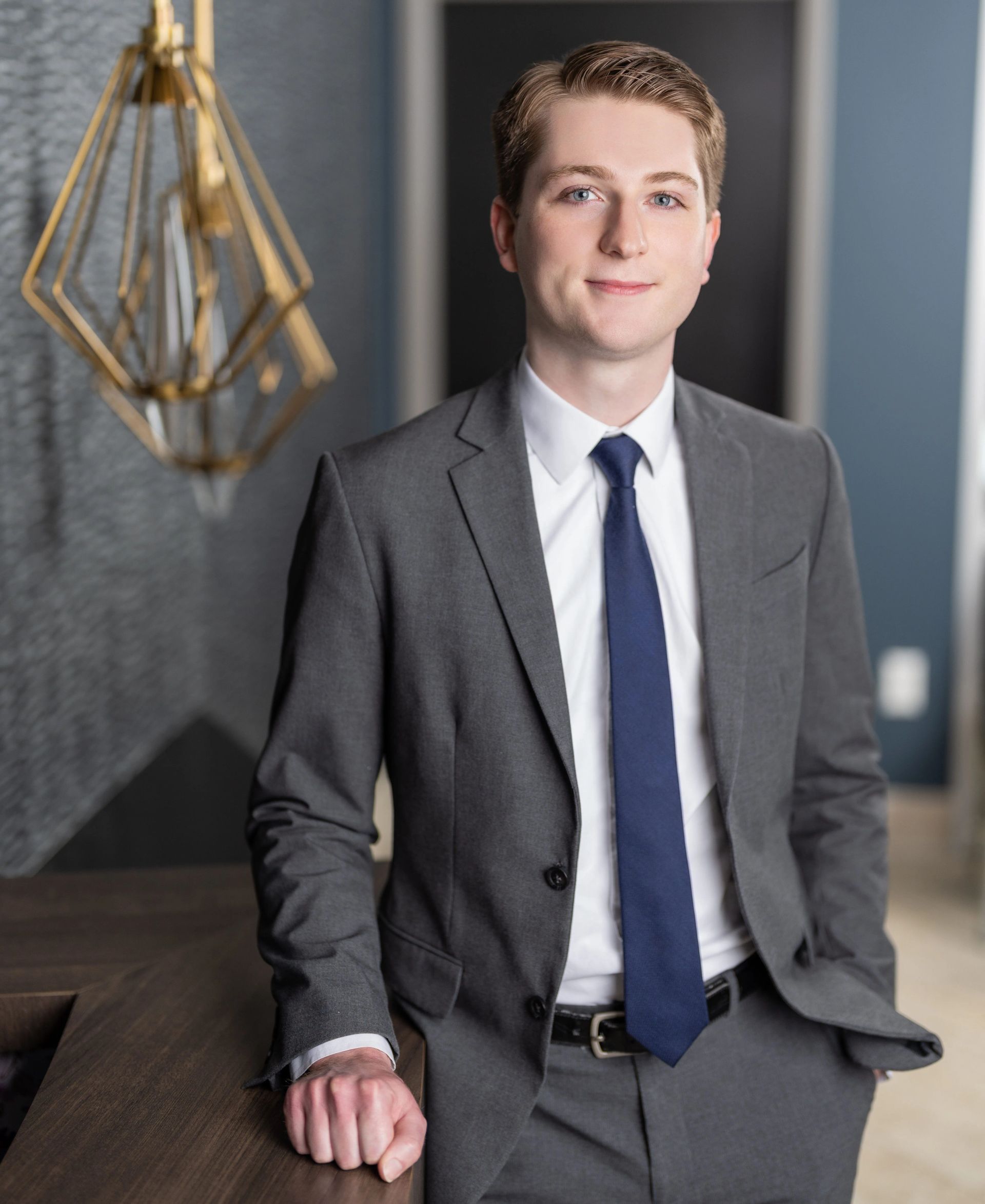 Man in gray suit with blue tie, leaning on a dark wooden surface, smiling in an office setting.