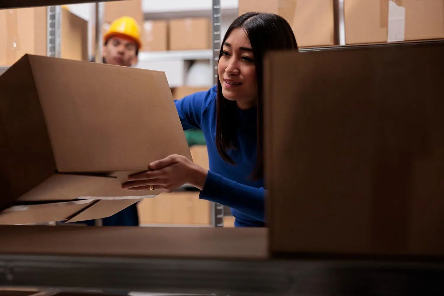Woman in blue shirt placing box on shelf in warehouse; worker in background.