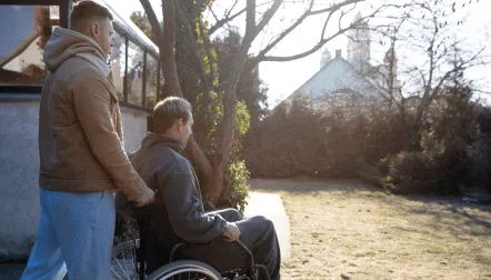 A person pushing a wheelchair with another person in it, outside a building with a white steeple.