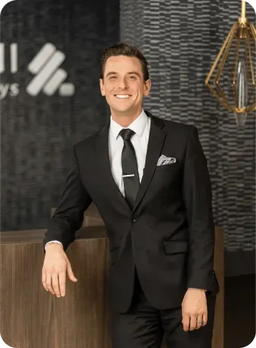Man in black suit smiles, leaning on a wooden counter with a logo, modern office setting.