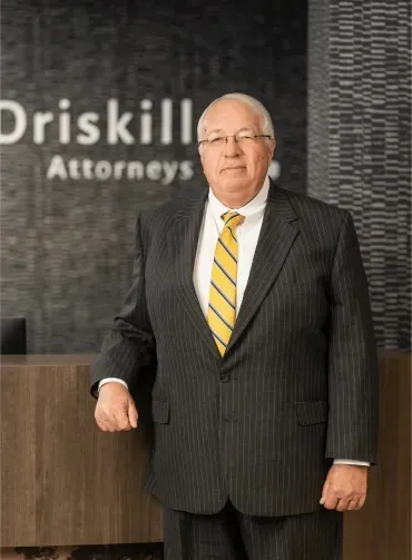 Man in suit standing in front of a law firm sign and reception desk. He wears glasses and a yellow tie.