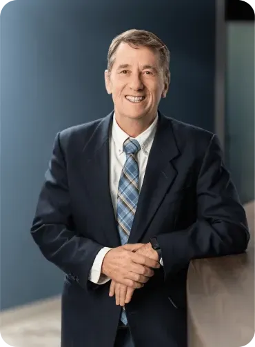 Man in a suit smiles, leaning on a counter against a blue wall.