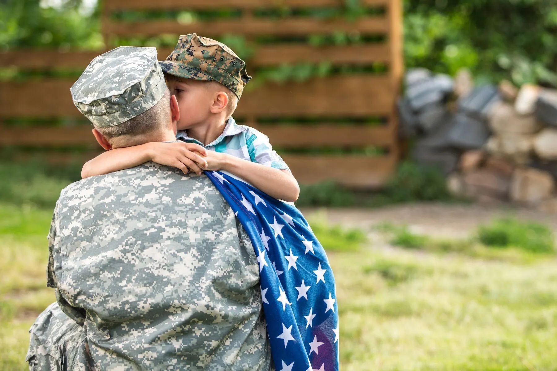 Soldier in camouflage uniform carries a child, who is wearing a camouflage hat and kissing the soldier, draped in an American flag.