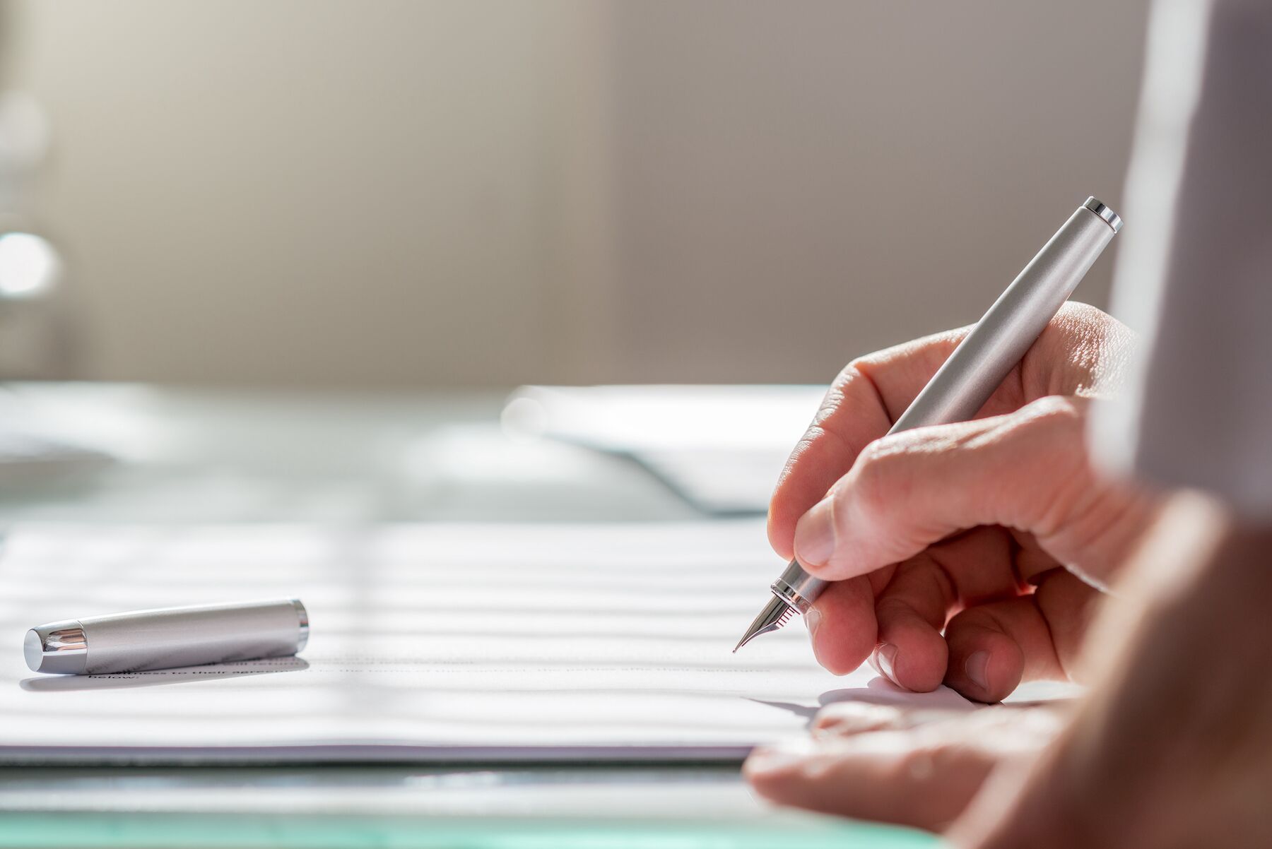 Hand writing on a paper with a silver pen, the pen cap lies on the desk in front of the writing.