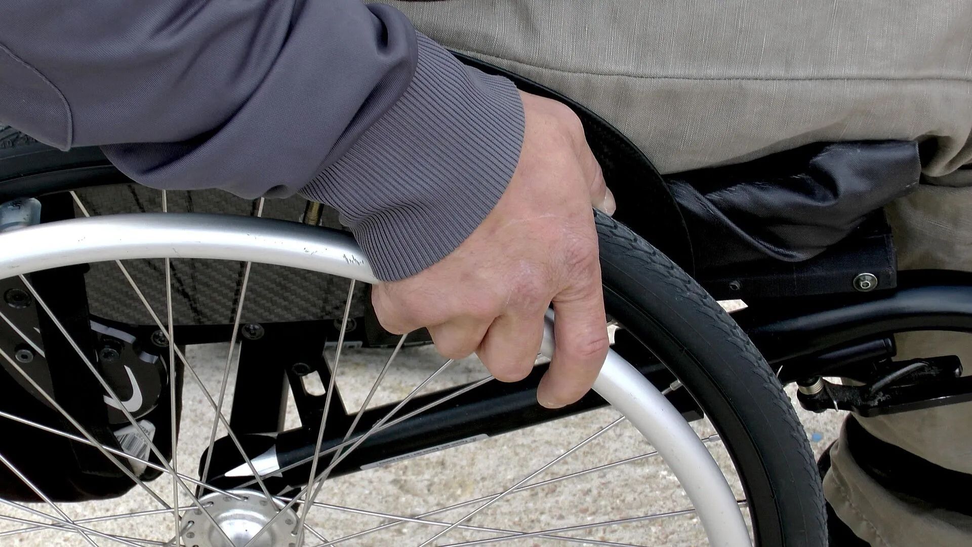 Person's hand on a wheelchair wheel, gripping the tire. Gray jacket sleeve and beige pants are visible.