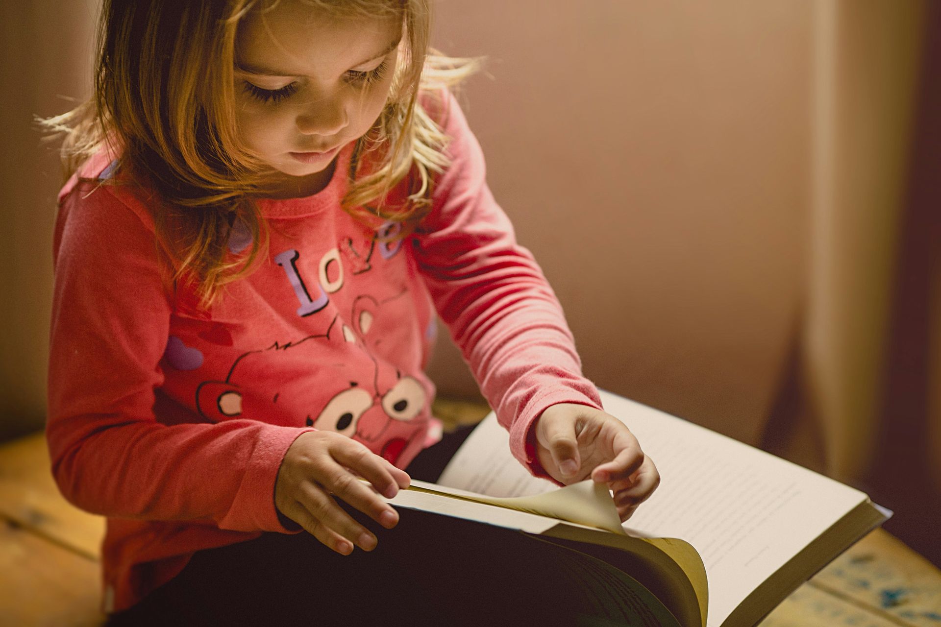 A little girl in a pink shirt is reading a book.