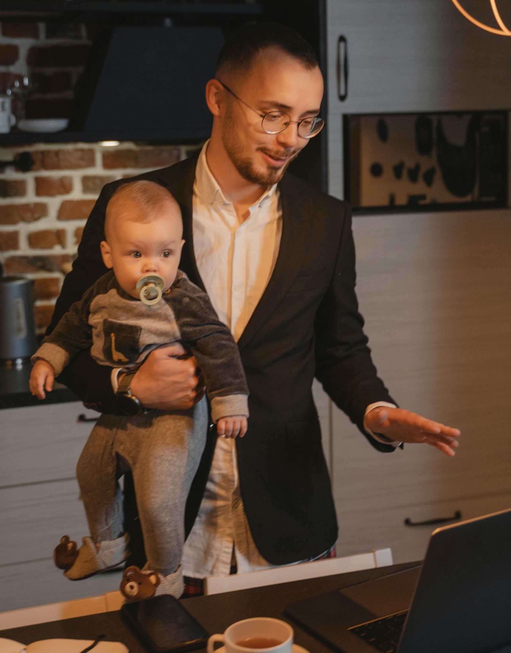 Man in a suit holds a baby with a pacifier while working on a laptop in a kitchen setting.