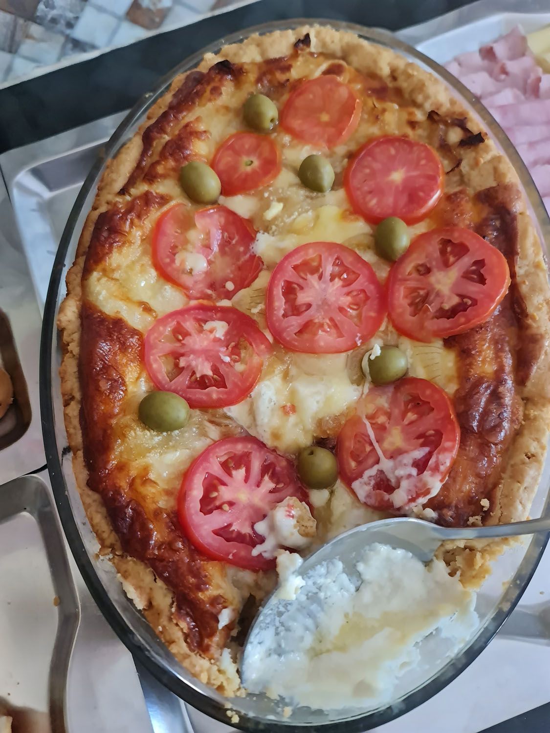 Uma pizza com tomates, azeitonas e queijo em um prato de vidro com uma colher dentro.
