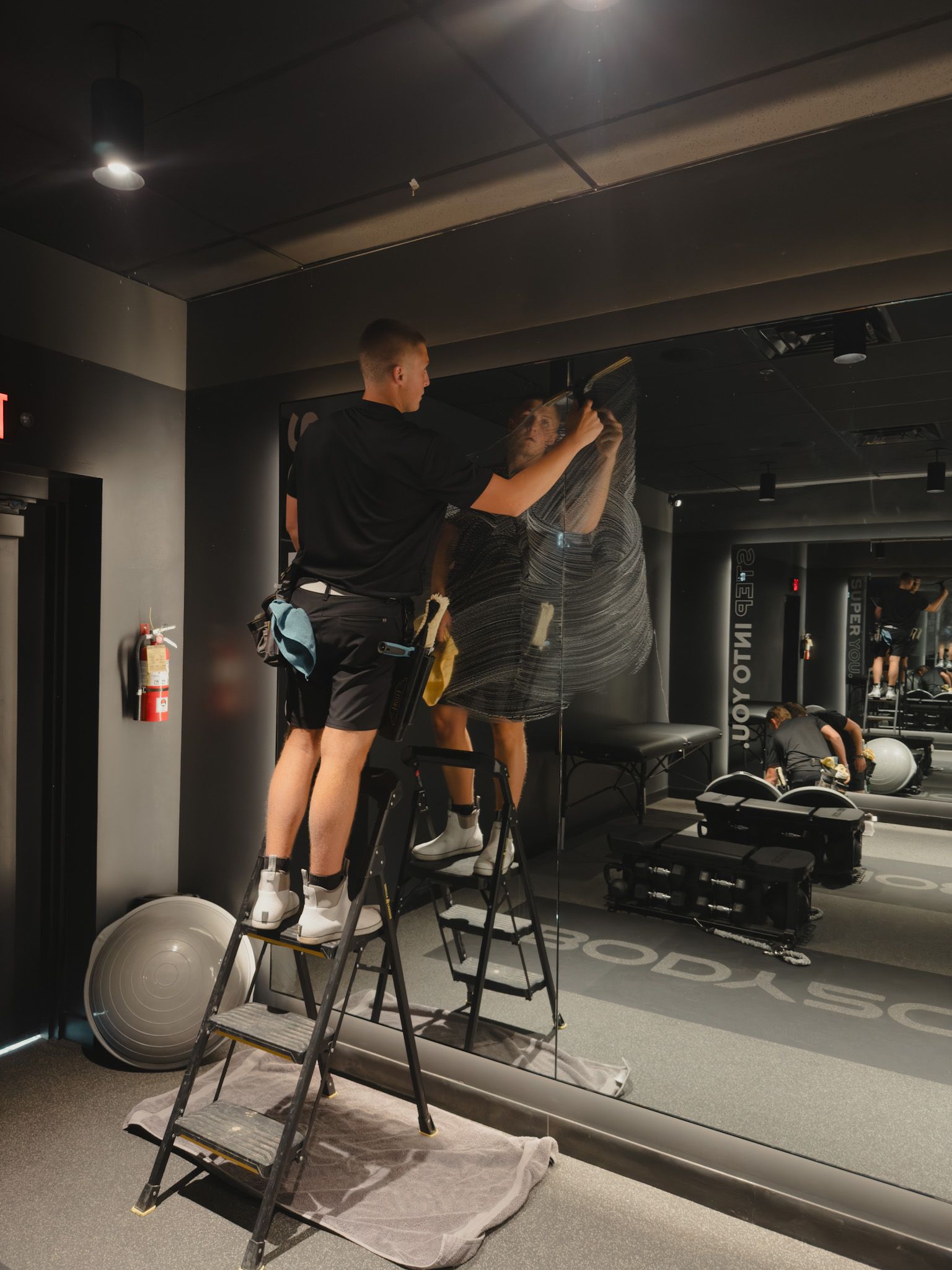 A man is standing on a ladder in a gym cleaning a window.
