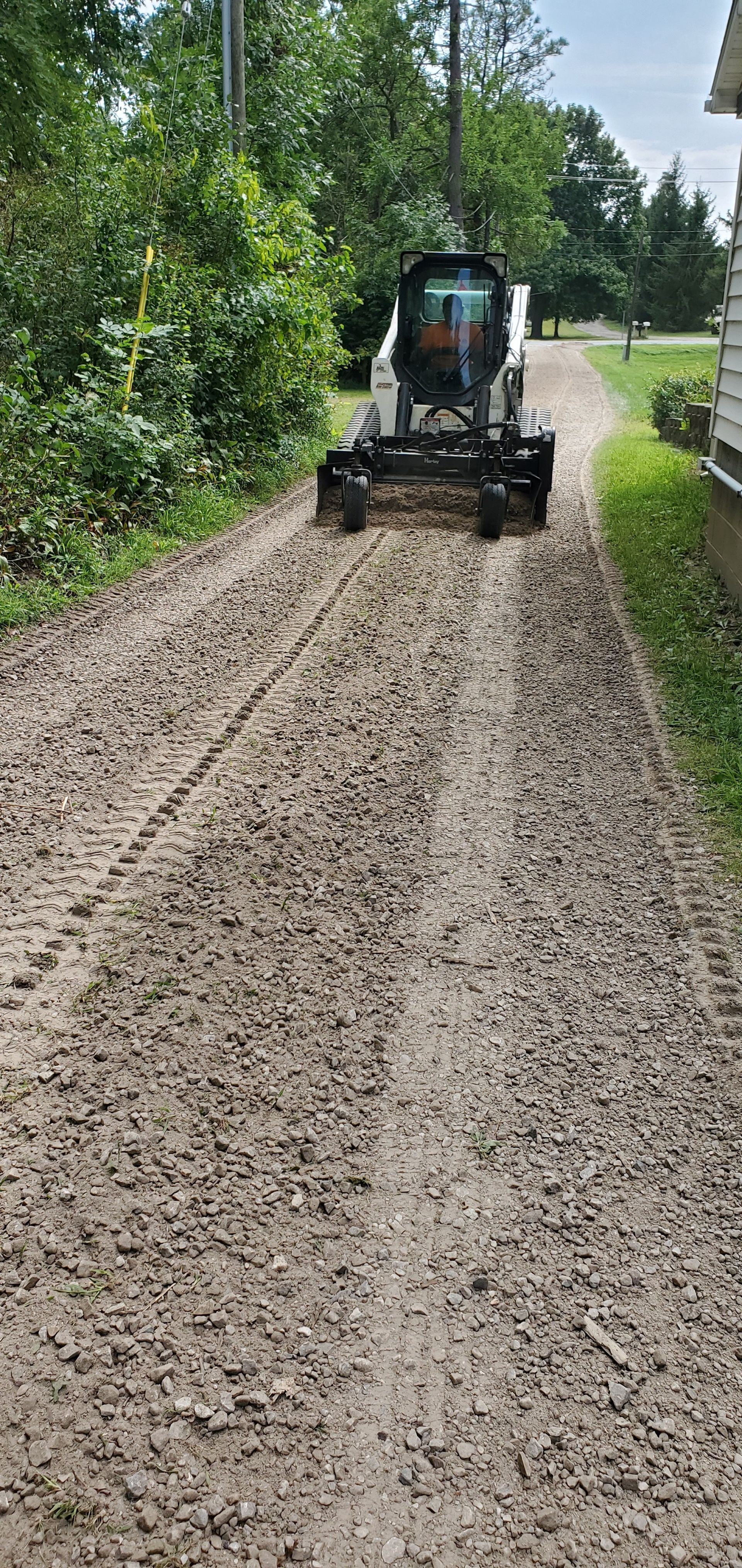 Bobcat grading a gravel driveway. Trees on either side, house on right.