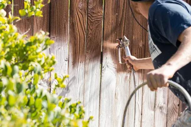 Person spray-painting a wooden fence outdoors with a paint sprayer and hose