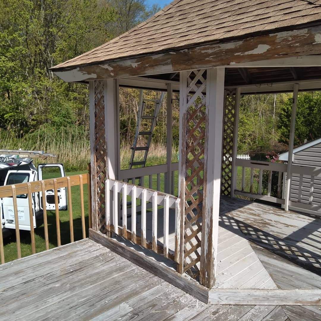 Weathered wooden gazebo with lattice posts and white railings on a sunny porch overlooking trees and parked cars