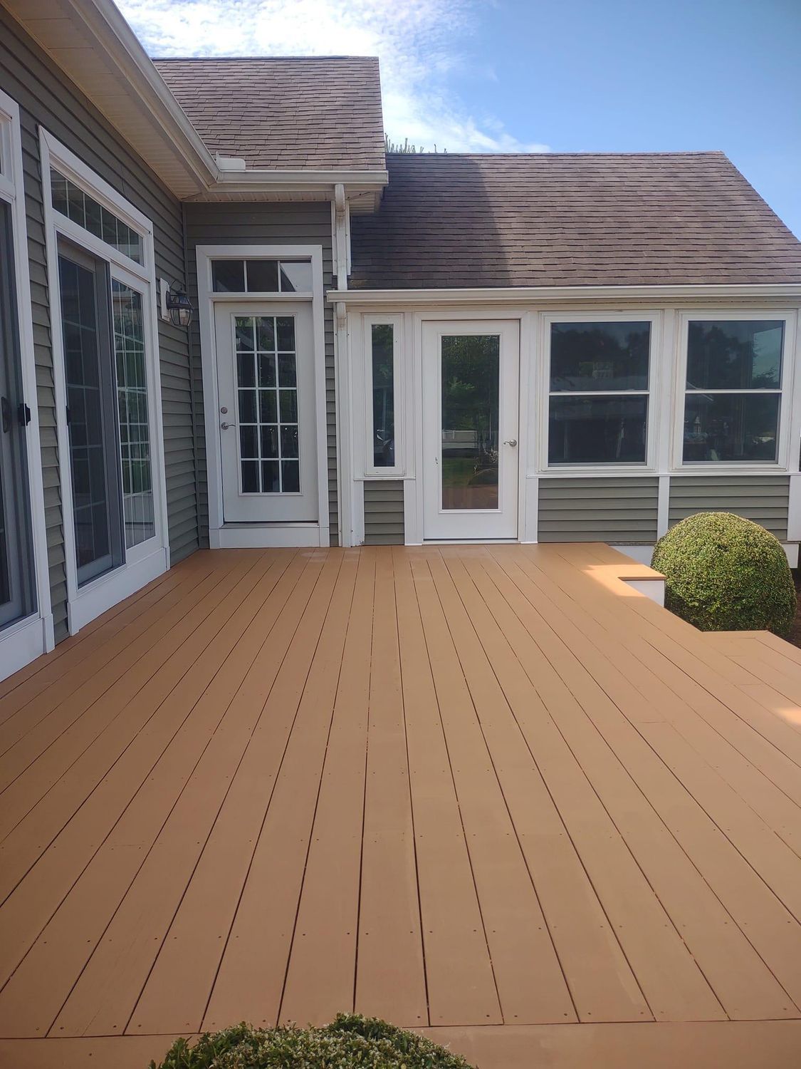 Empty tan wooden deck outside a house with white siding and windows, under a blue sky