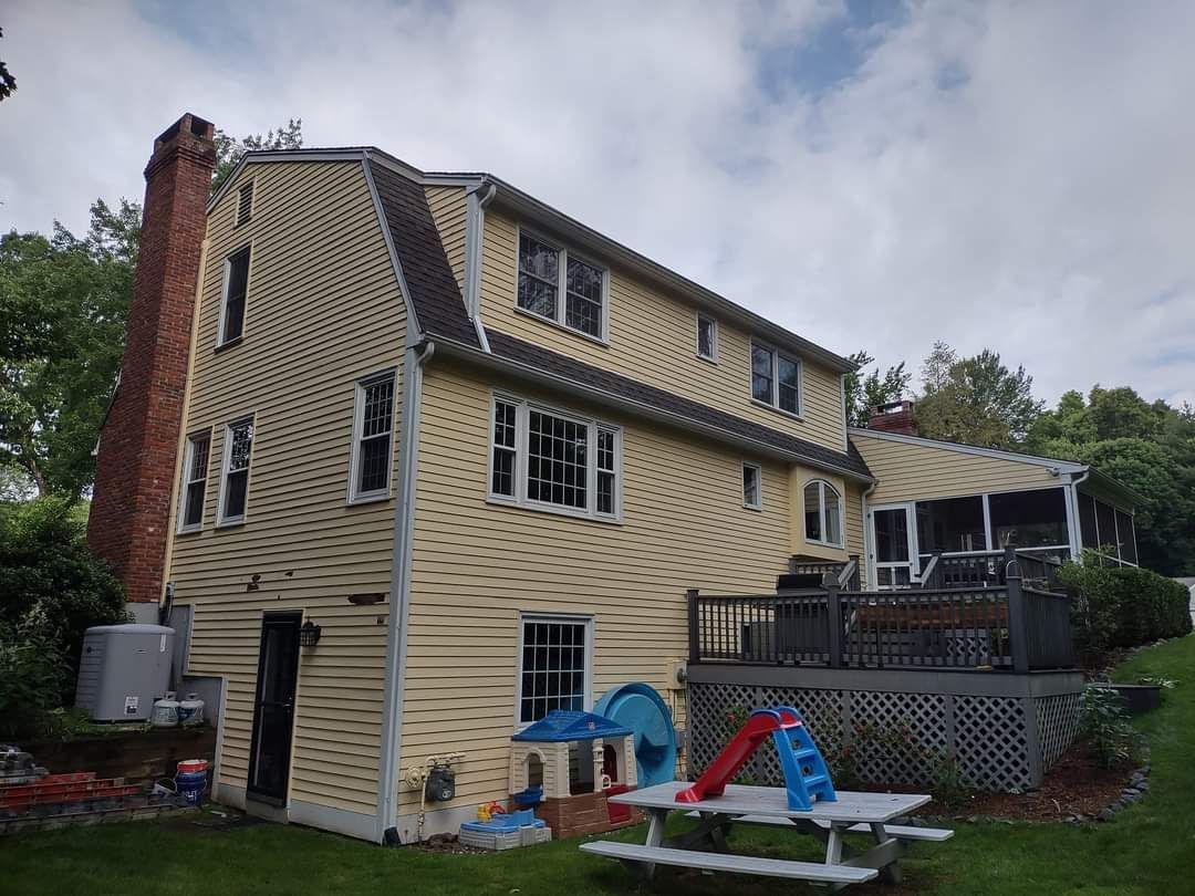 Damaged beige house with cracked siding, red-brick chimney, and a backyard playset under cloudy sky