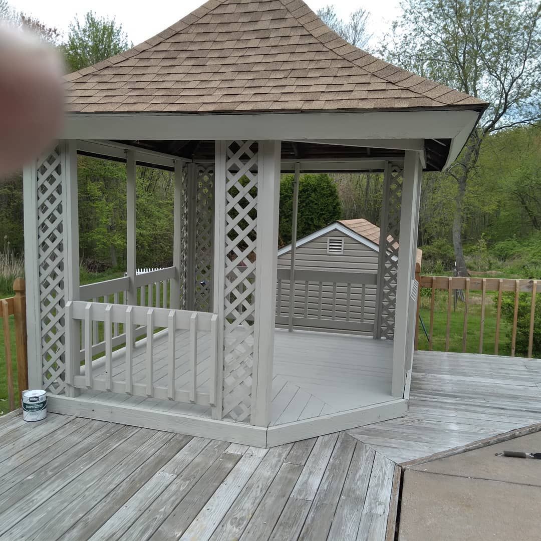 Wooden gazebo with lattice sides on a deck, viewed outdoors in a wooded yard.