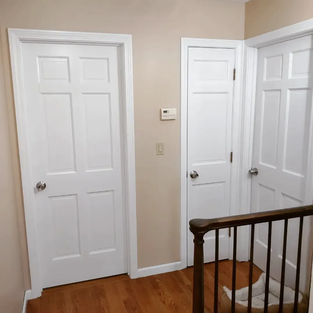 Hallway with three white doors, beige walls, hardwood floor, and a stair railing in the foreground