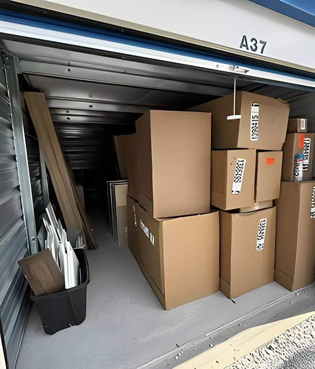 Storage unit filled with cardboard boxes and some long objects. A black bin sits near the door.