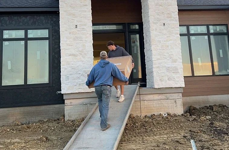 Two men carrying a large box up a concrete ramp into a building under construction.