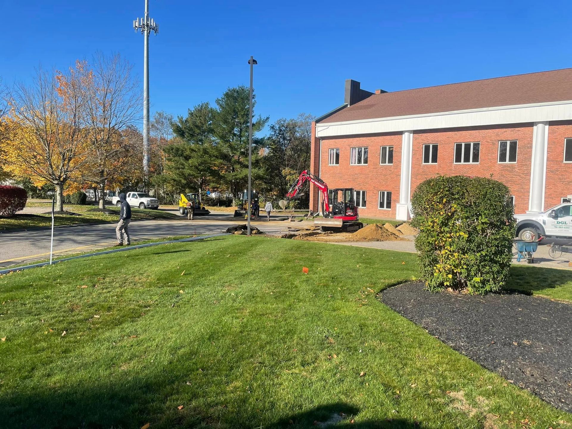 A red mini excavator works on a small construction project near a brick building on a sunny day.