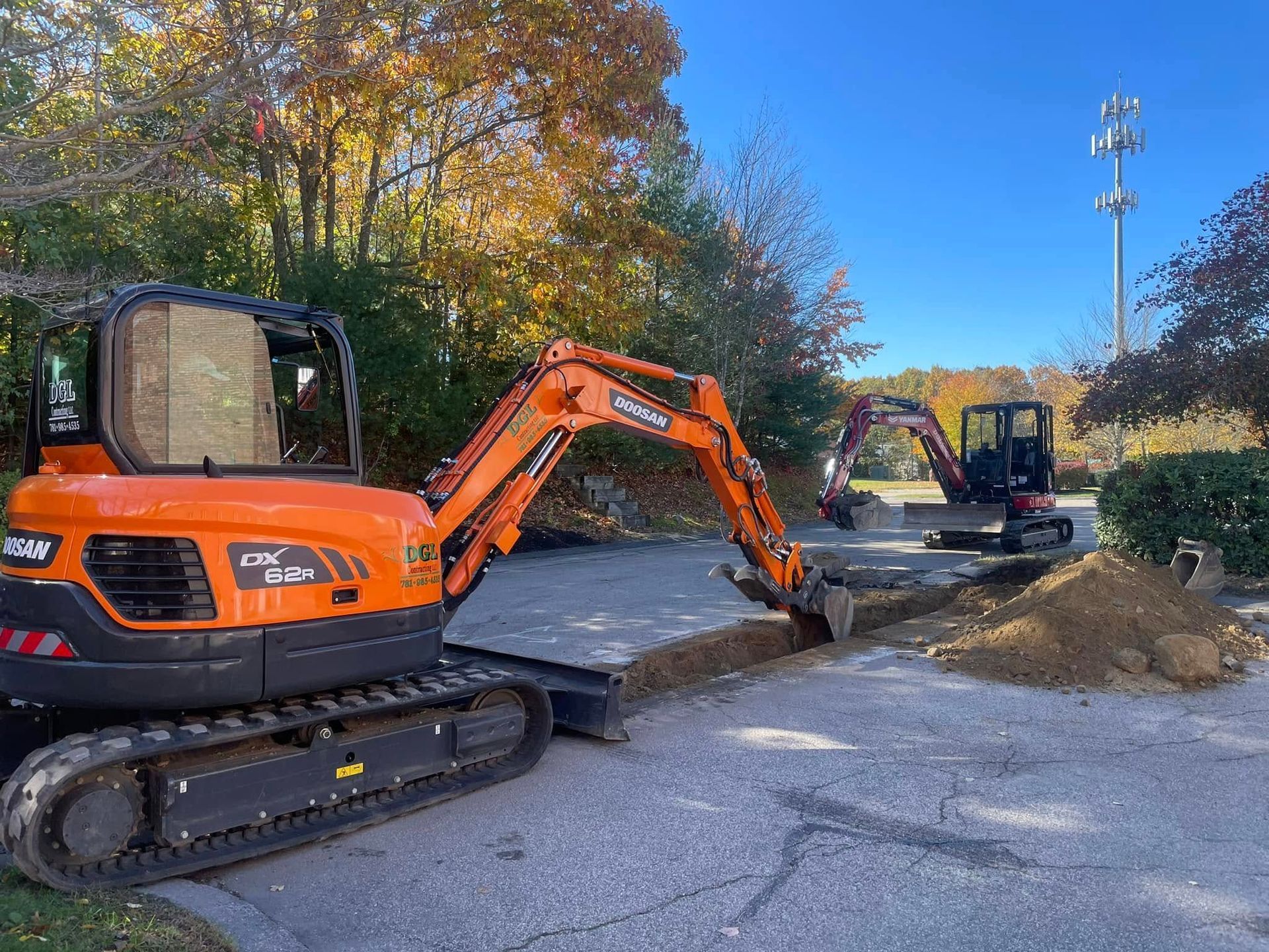 Two orange excavators work on a dirt trench in a paved area, with trees and a cell tower in the background.