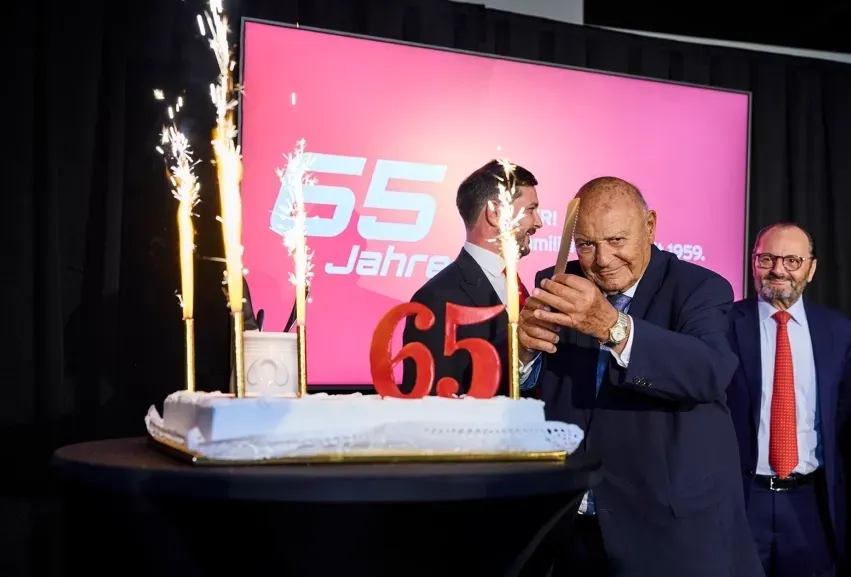 Ein Mann schneidet bei einer Veranstaltung mit Wunderkerzen, rotem Hintergrund und umstehenden Kollegen eine Torte an.