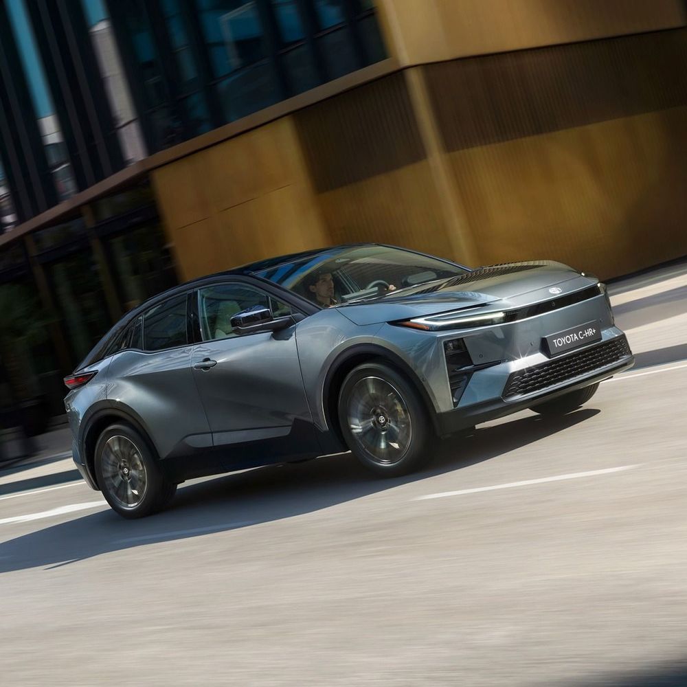 Ein metallicgrauer Toyota C-HR fährt auf einer Straße in der Nähe eines modernen, goldfarbenen Gebäudes.