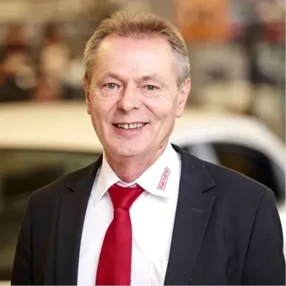 Ein Mann im Anzug und mit roter Krawatte lächelt vor dem verschwommenen Hintergrund eines Autohauses.