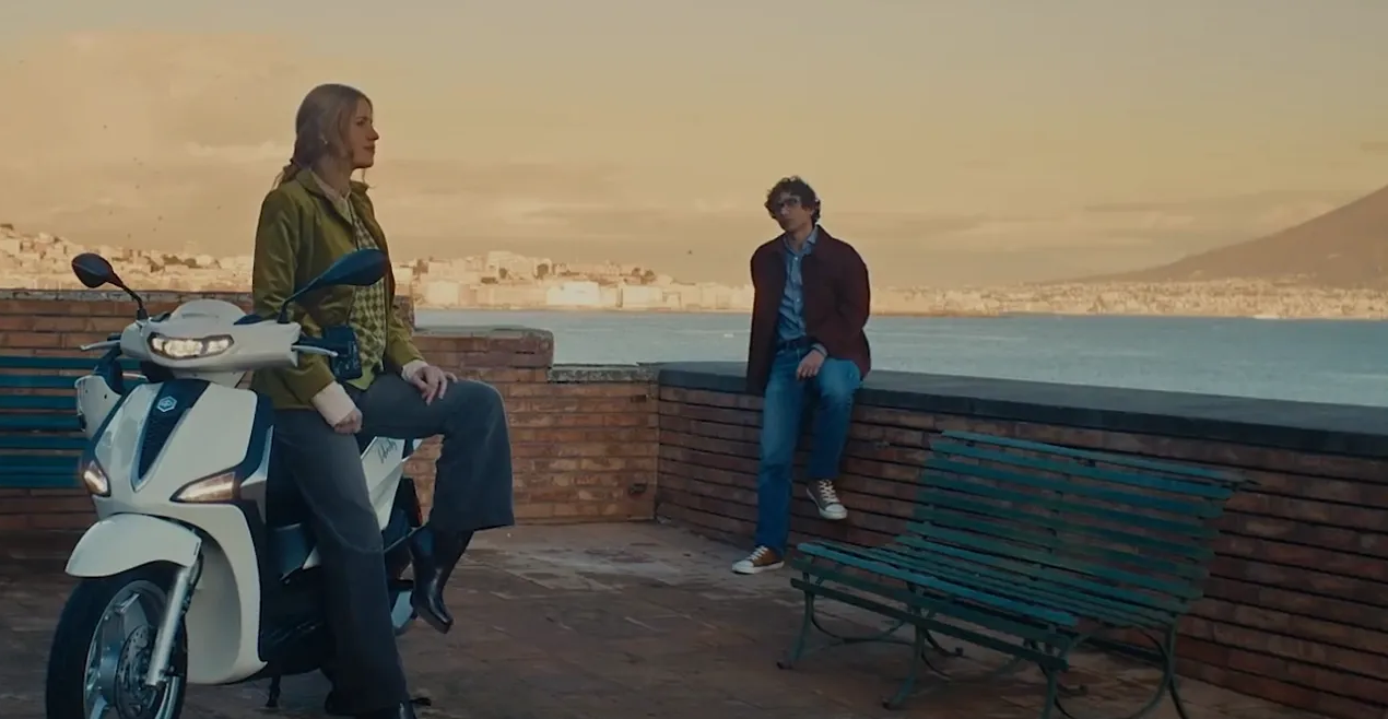 Eine Person sitzt auf einem weißen Roller, während eine andere an einer niedrigen Backsteinmauer lehnt