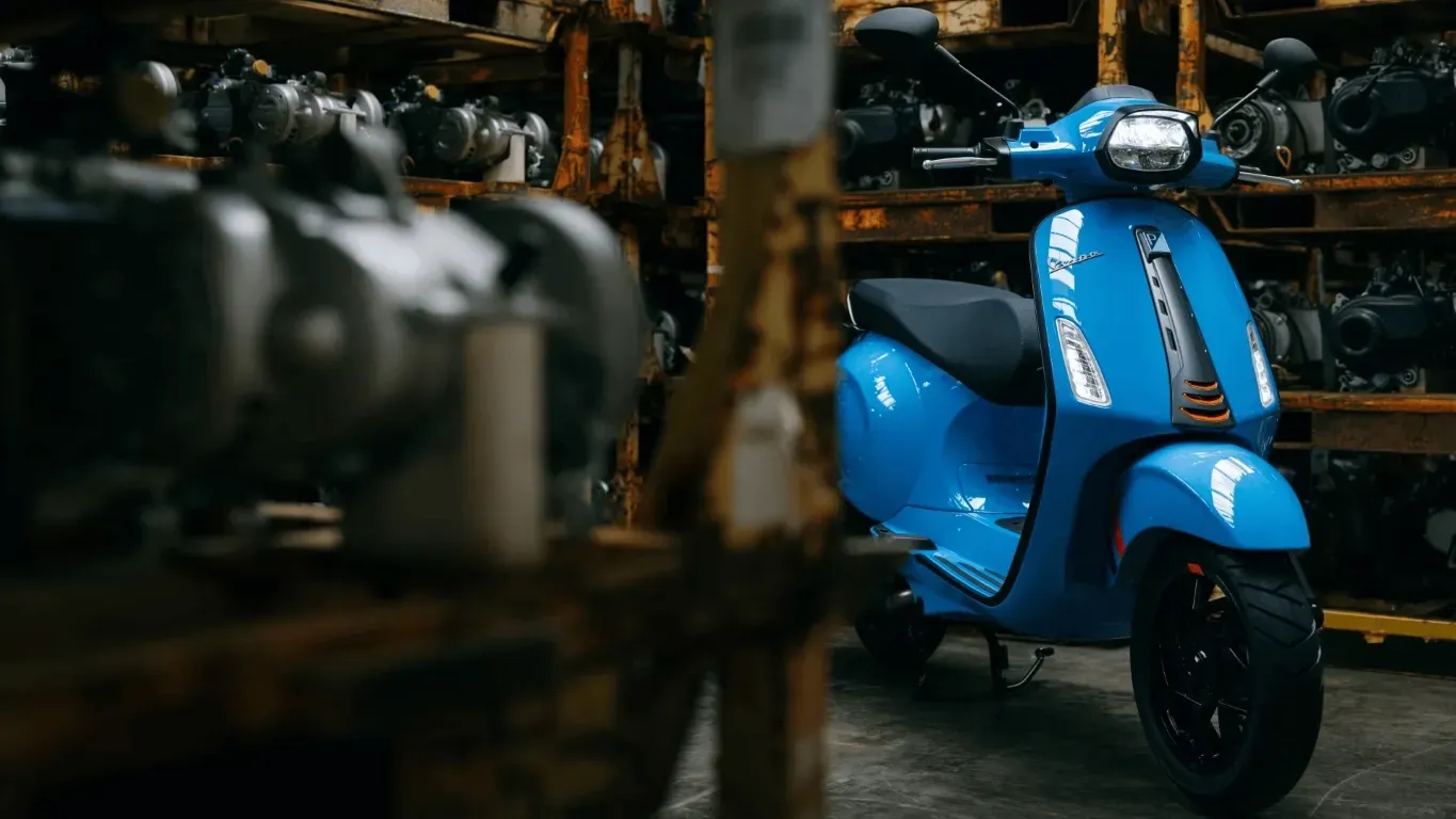 Ein leuchtend blauer Vespa-Roller parkte in einer düsteren Industriehalle voller Regale mit Maschinenteilen.