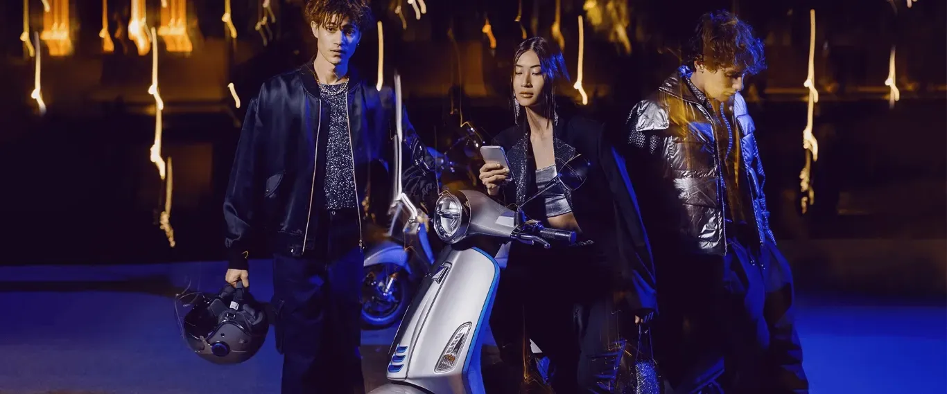 Drei Personen in stilvoller, dunkler Kleidung stehen mit einem hellen Roller vor einem dunklen Hintergrund