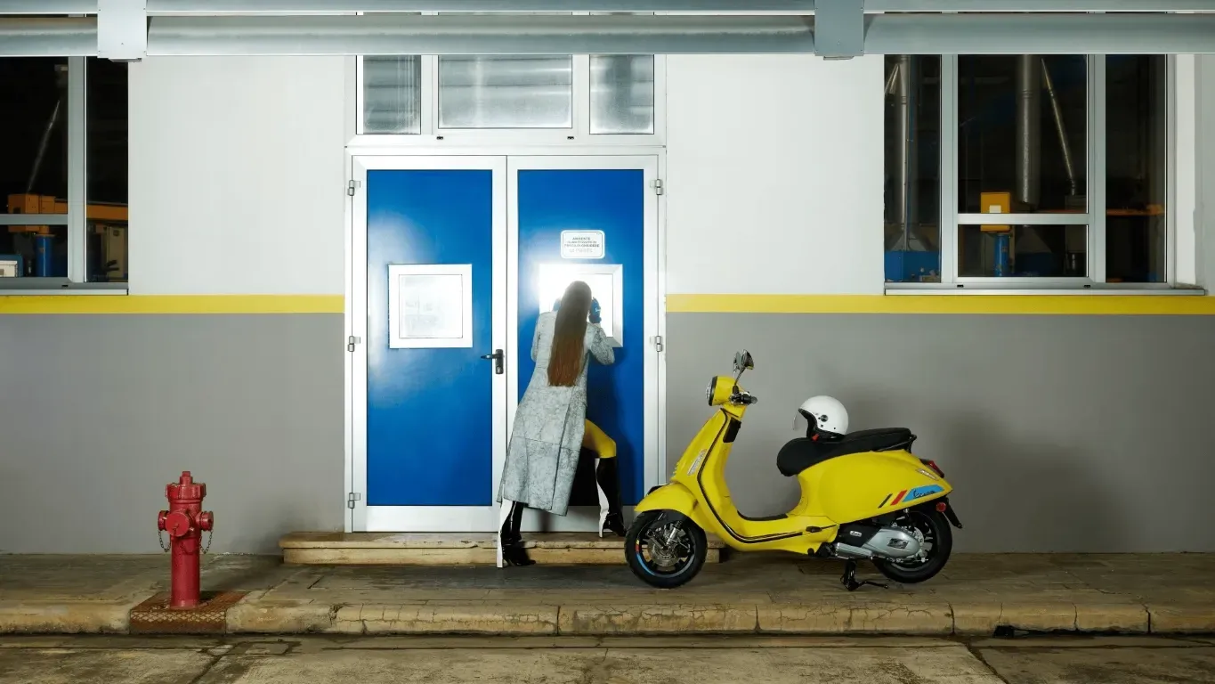 Eine Person in einem langen Mantel steht vor einer hellblauen Doppeltür, neben einem geparkten gelben Roller