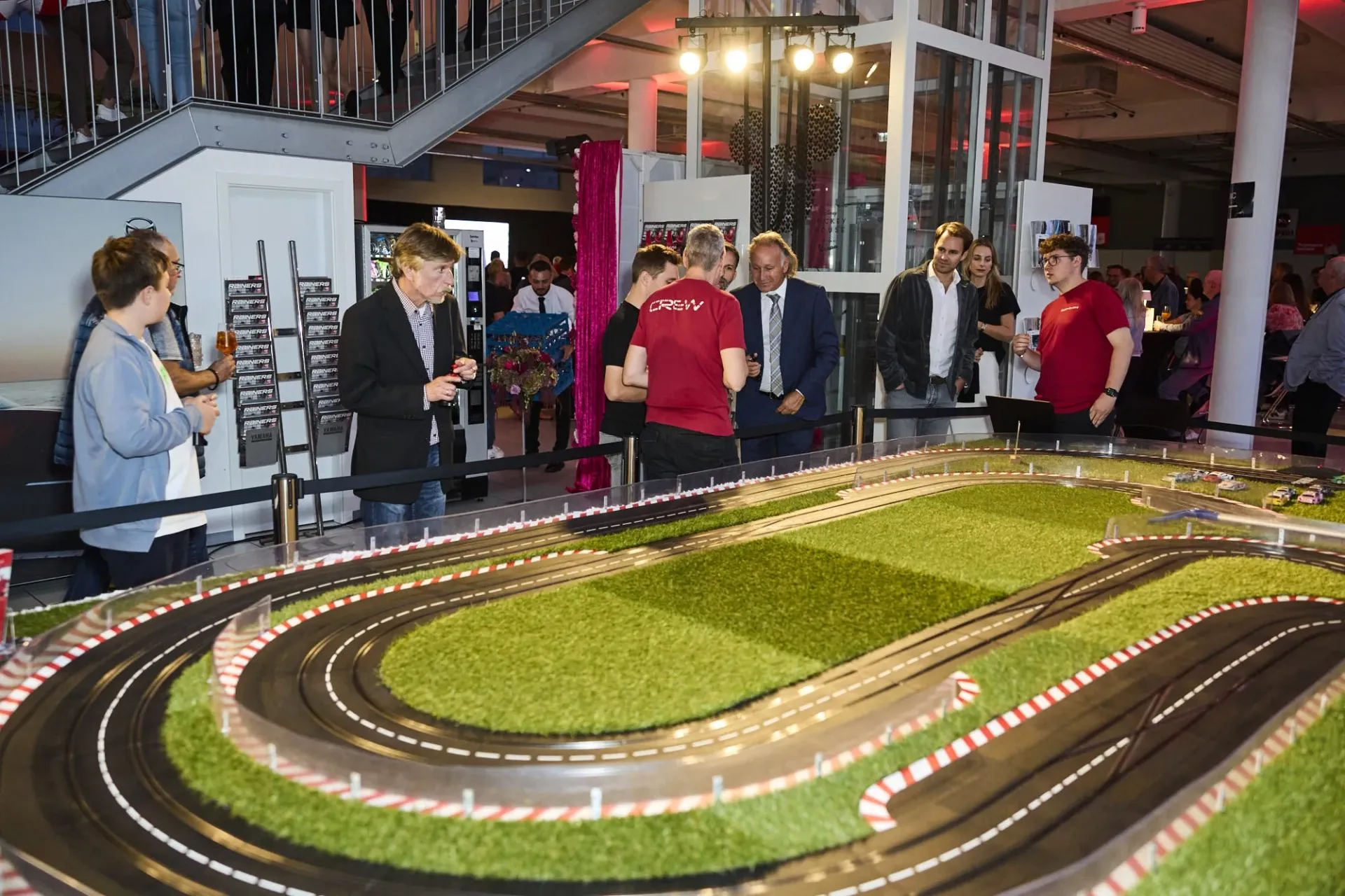 Eine große Slotcar-Rennbahn in einem hellen, modernen Veranstaltungsraum, um die sich mehrere Personen versammelt haben.