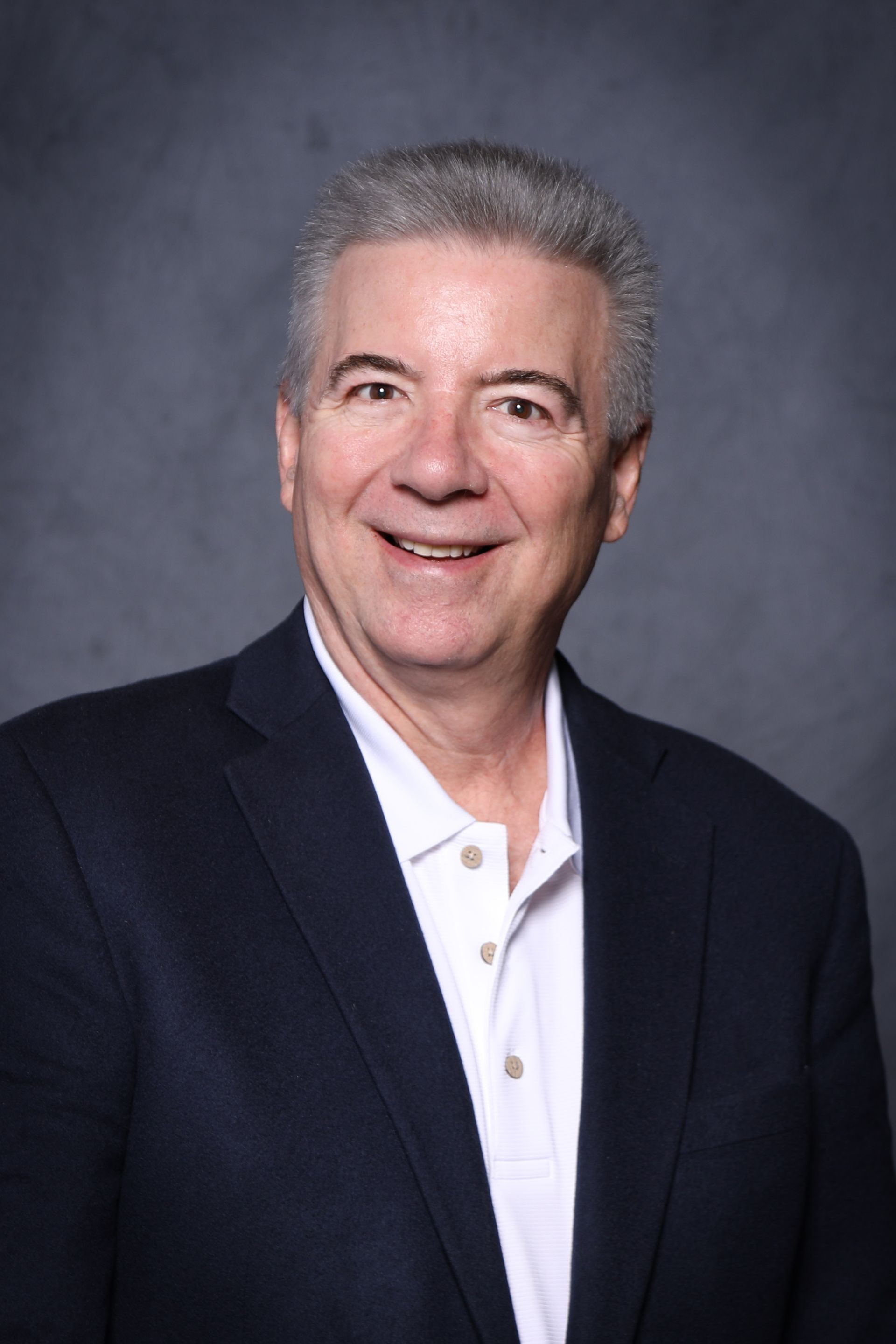 Smiling man in a navy blazer and white polo shirt against a gray backdrop.