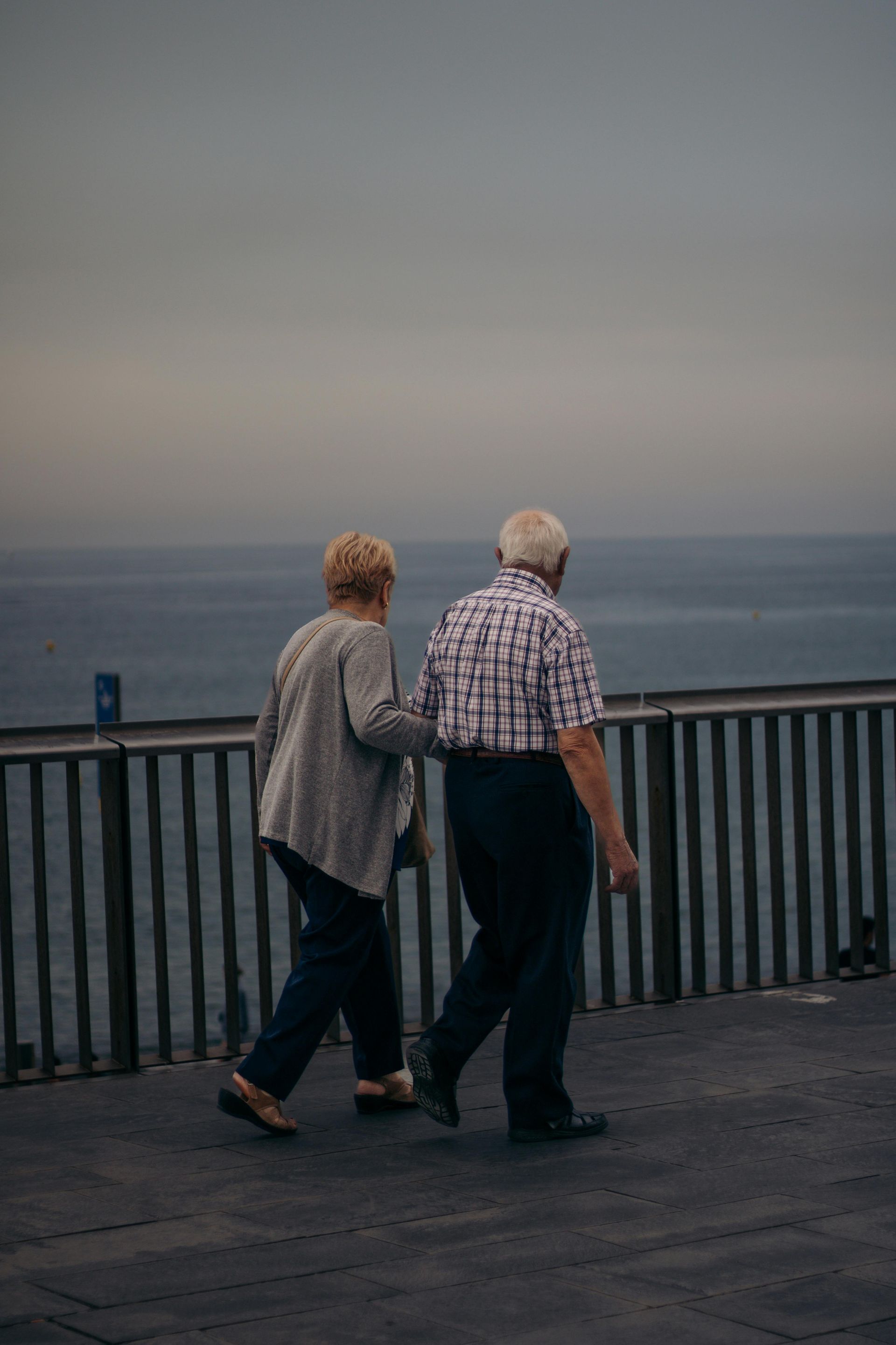 Elderly couple walking together near a body of water, gray sky.