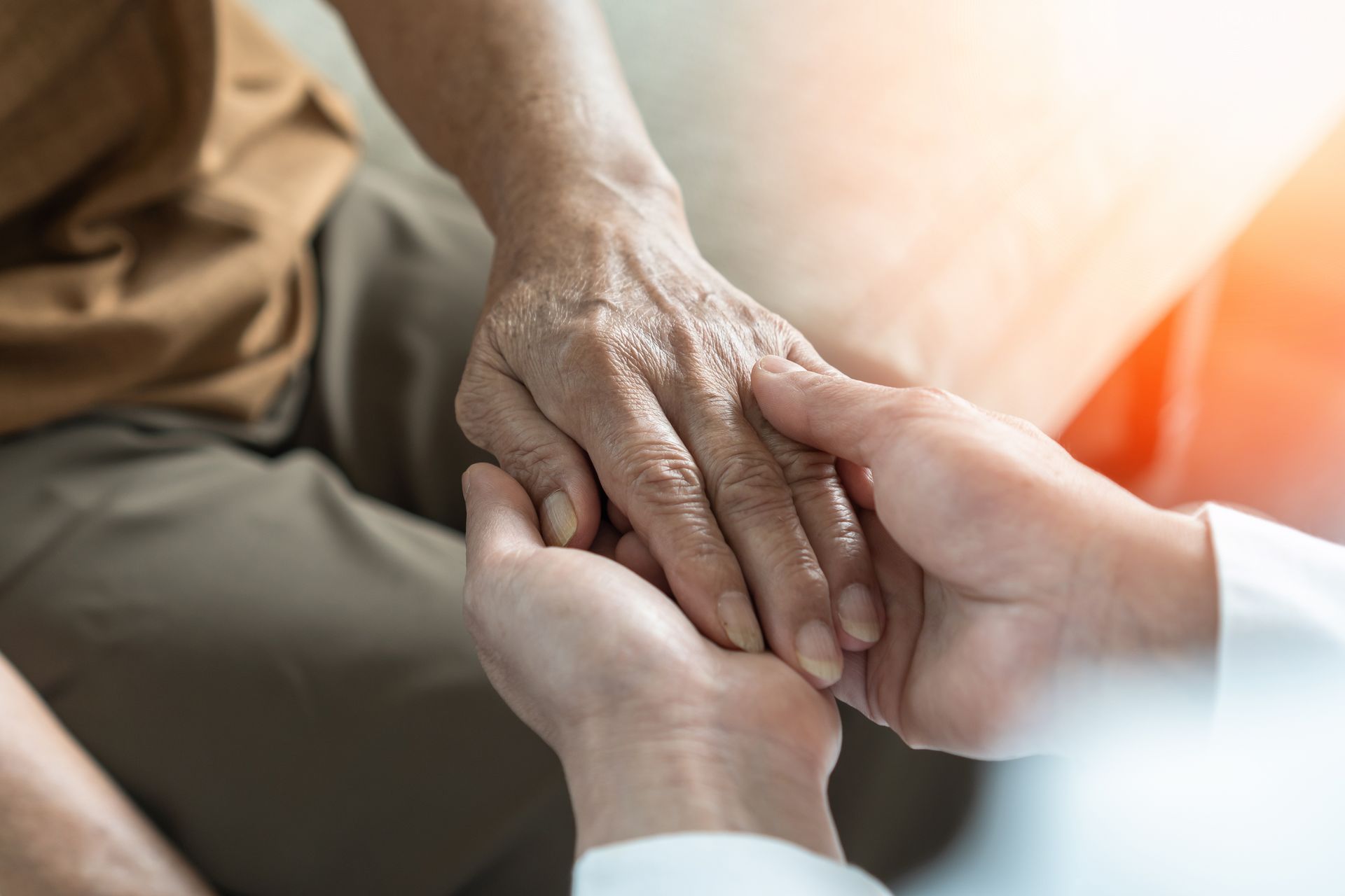 Two hands clasped: Elderly person's hand held by someone wearing a white coat, offering comfort.