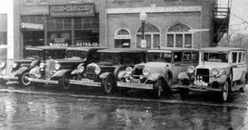 A black and white photo of a row of old cars parked in front of a building.