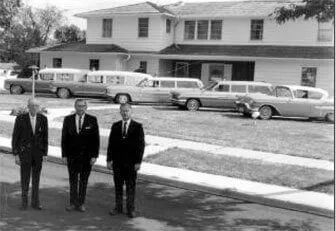 Three men are standing in front of a house with a row of cars parked in front of it.