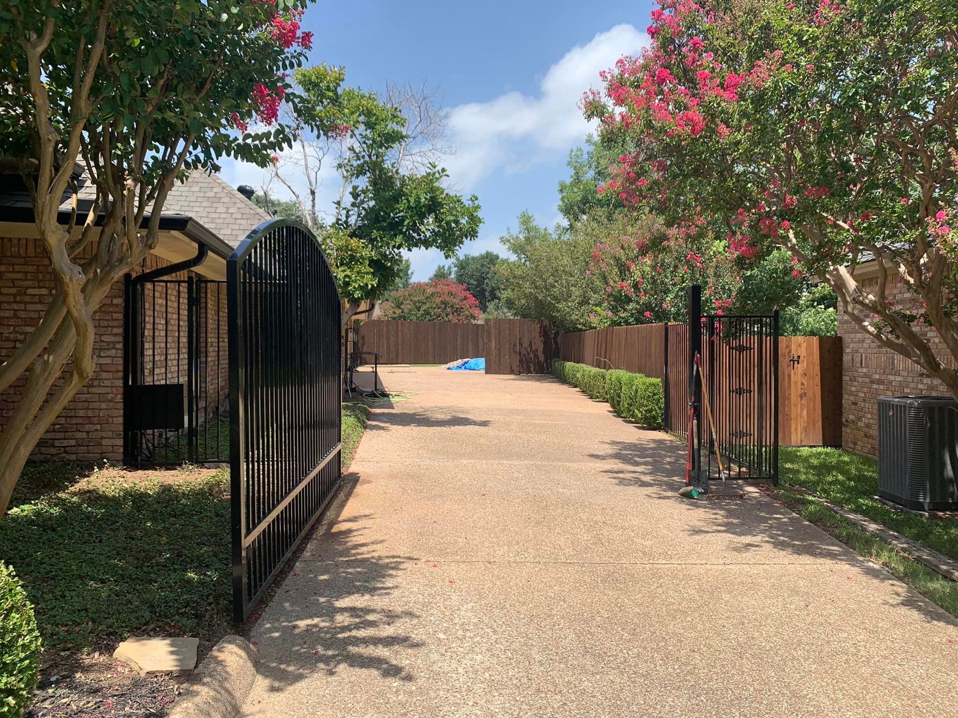 Black gates open to a gravel driveway lined with trees and hedges on a sunny day.