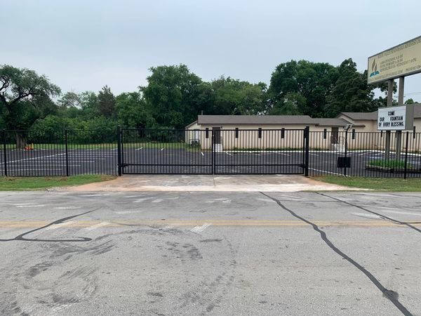 Black metal gate and fence enclosing a parking lot in front of a one-story building; overcast sky.