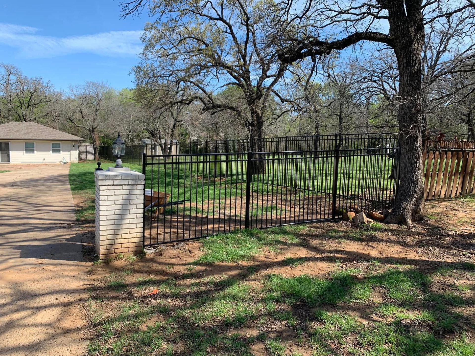 Black metal fence encloses a yard with trees and green grass; a brick pillar stands at the driveway entrance.