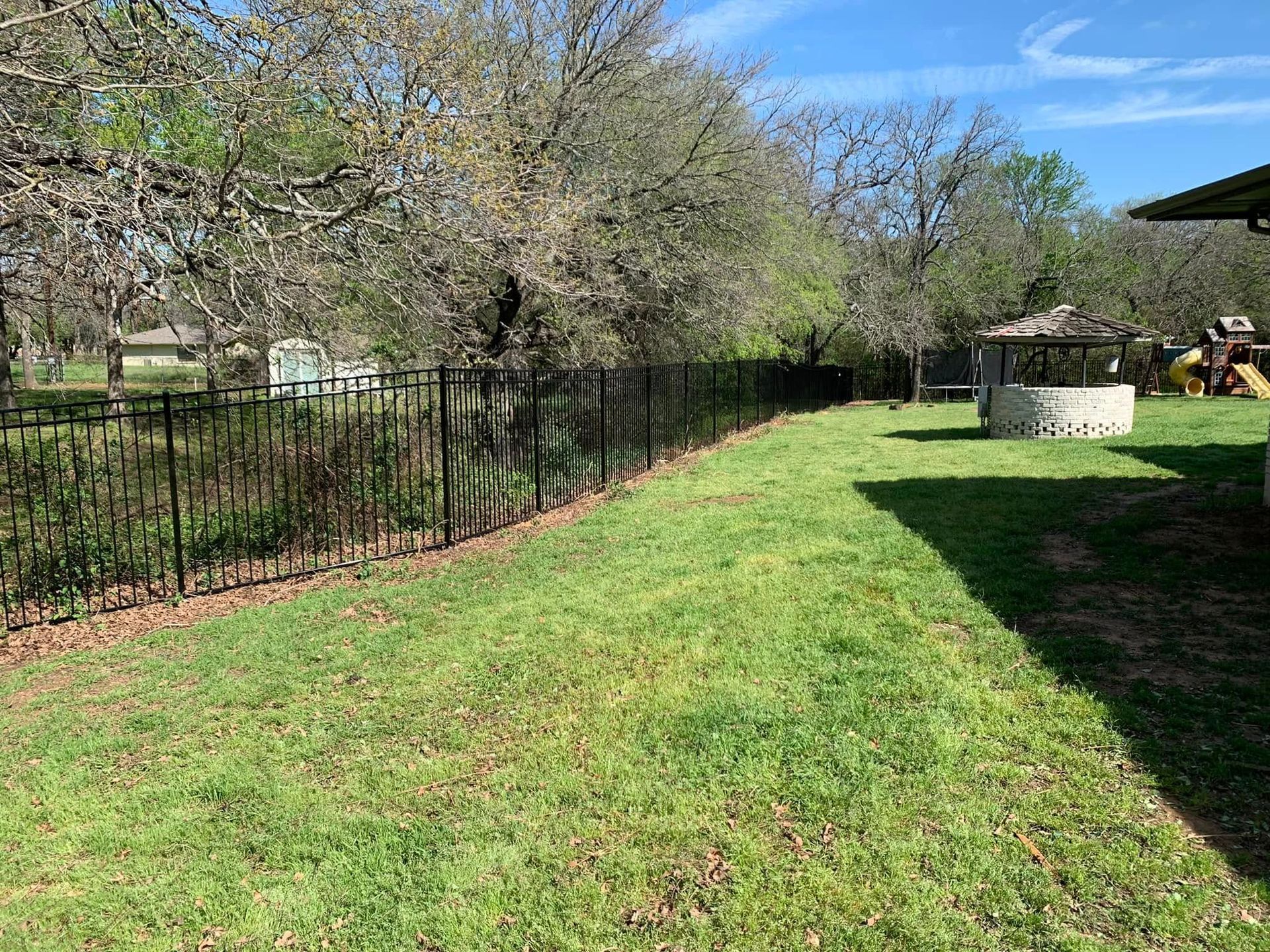 Black fence along a grassy yard with trees, a gazebo, and a clear blue sky.