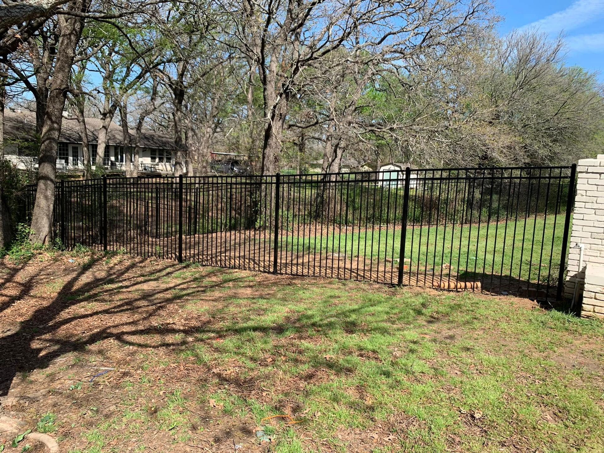 Black metal fence in a yard with trees, grass, and a stone wall.