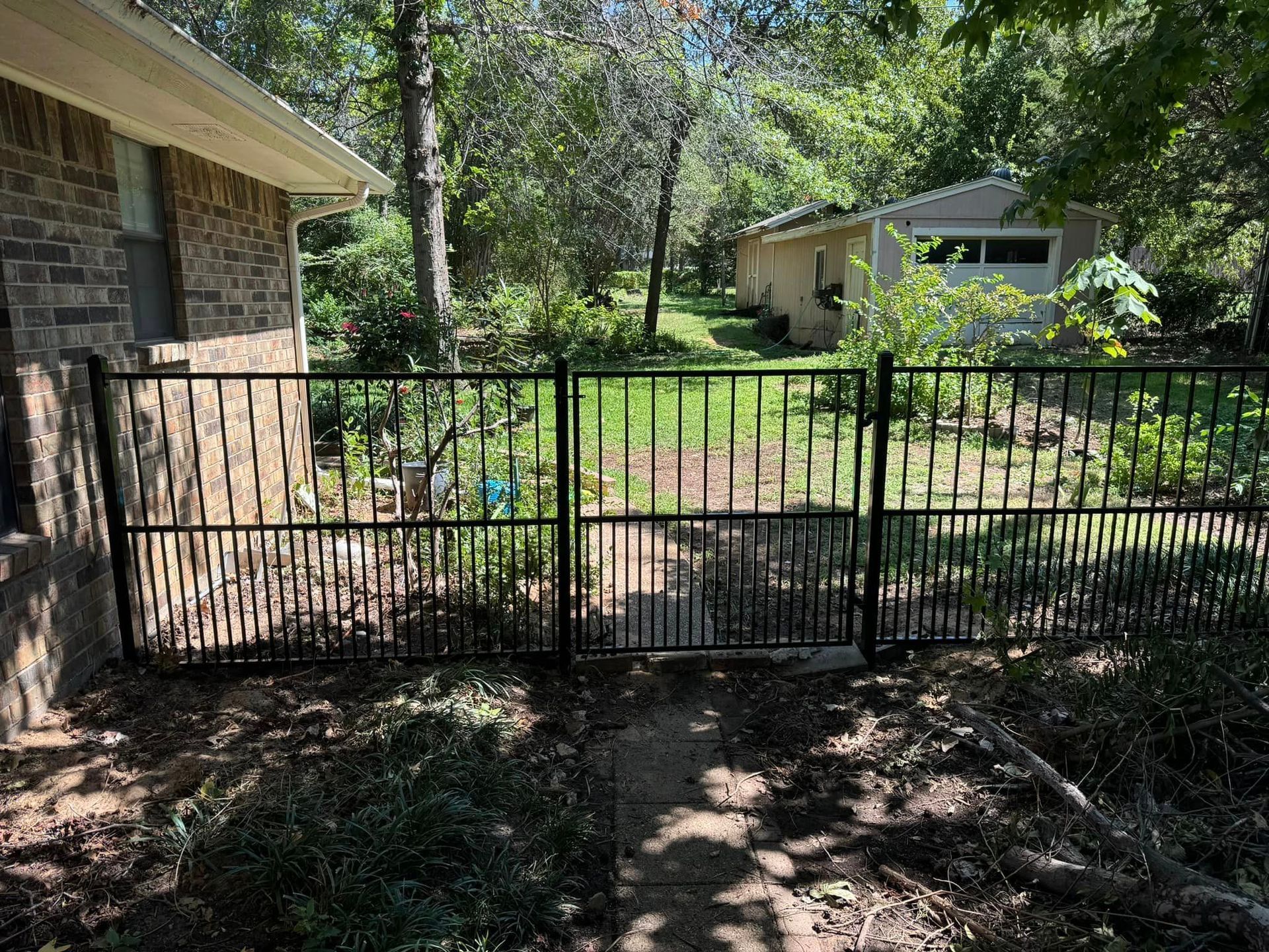 Black metal fence encloses a backyard path, near a brick house and trees.