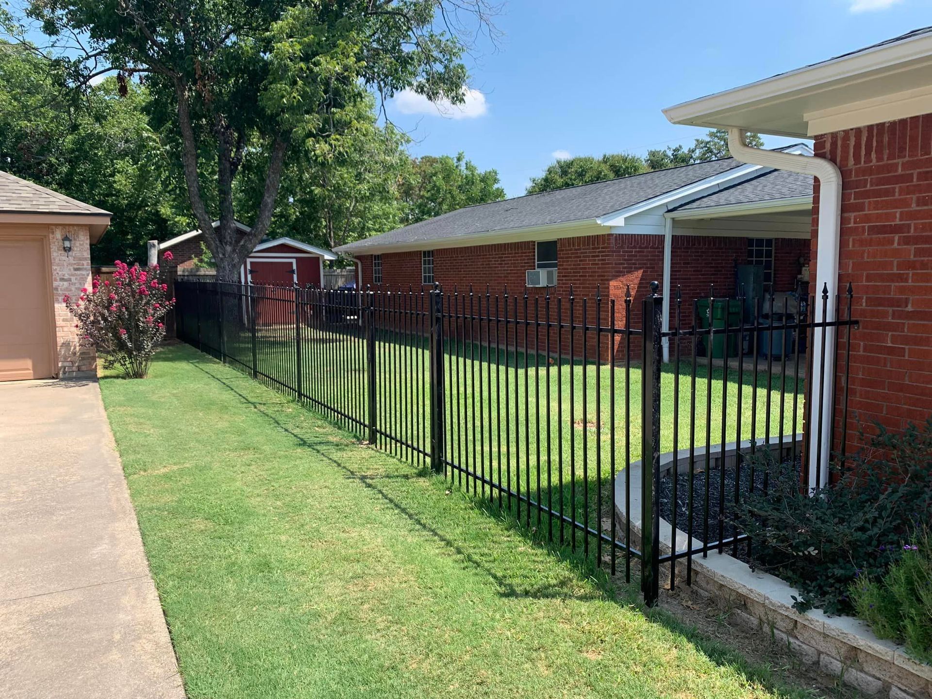 Black metal fence bordering a grassy yard next to a brick house on a sunny day.
